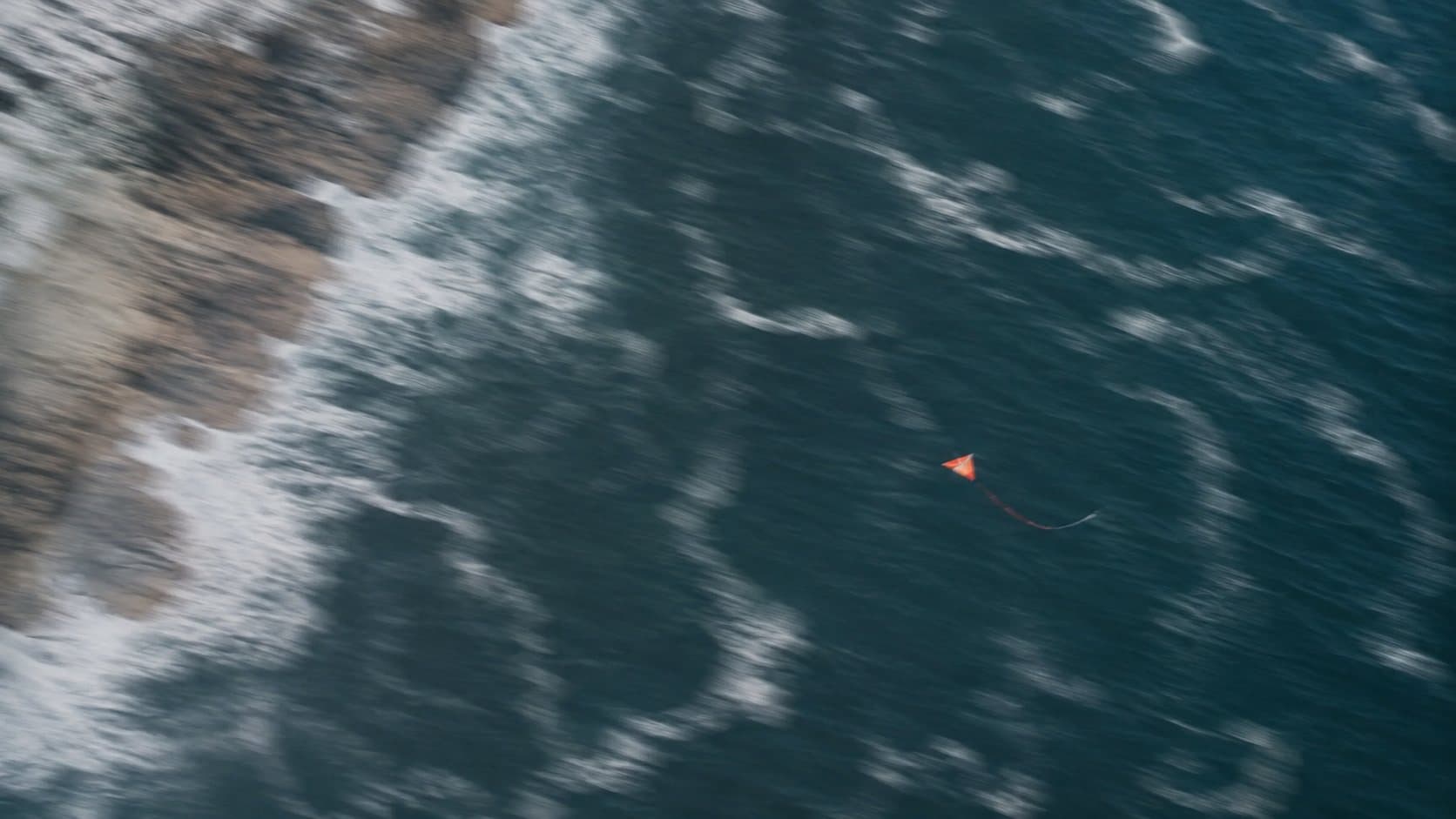 A lone orange kite gliding over a deep blue-green ocean beside a rugged, foam-laced coastline.